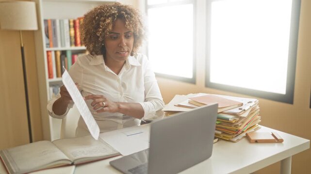 Woman flips documents and reviews notes at a desk with laptop, open book and stacked folders in an office building; concentration productivity.