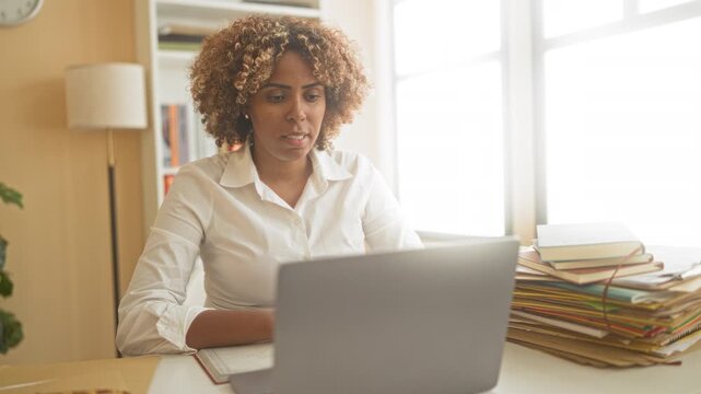 Woman pumps fists at laptop, showing hands and smiling at desk with stacked books and files in building; achievement joy.