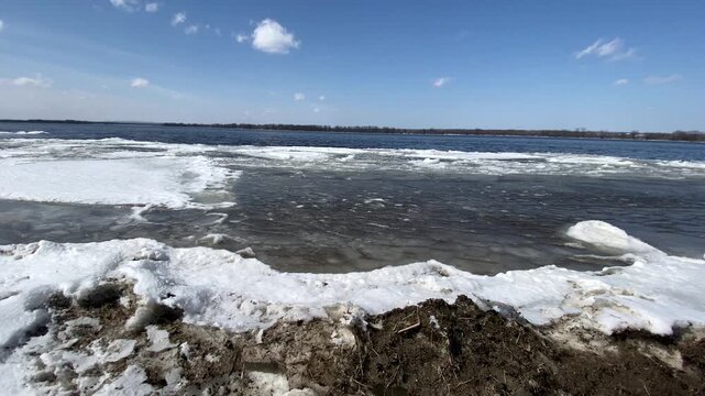 Melting ice floes and snow piled along a shoreline under a clear blue sky, bordering open dark blue river water during early spring thaw.