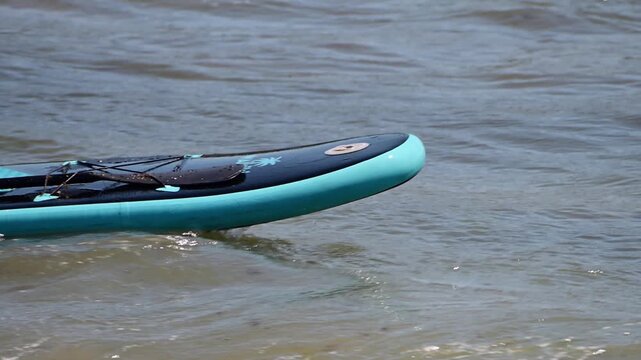A teal and dark blue stand-up paddleboard bobs gently in the shallow, rippling waves near a beach.