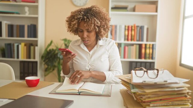 Woman holding smartphone and flipping book pages at office building desk with stack of papers, glasses and red cup; concentration productivity.