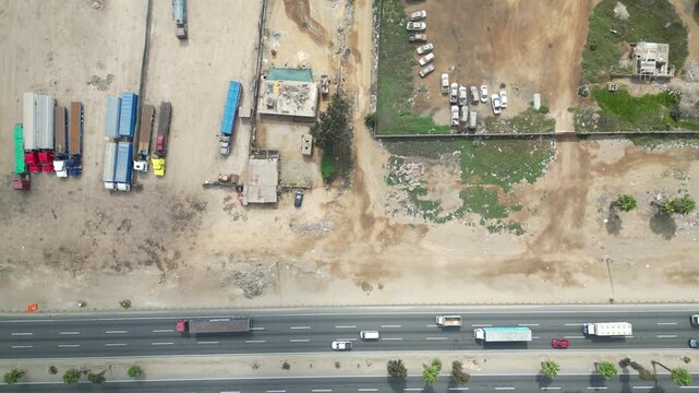 Aerial drone view of an abandoned vehicle scrapyard and parked semi-trucks along a dusty desert highway.