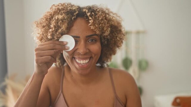 Woman holding a cotton pad to one eye while smiling with bra strap visible in a bright bedroom during a skincare routine; self care happy.