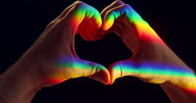 Close-up studio video of two hands forming a heart shape illuminated by rainbow-colored sunlight against a black background. Symbolic LGBTQ+ Pride concept representing love, equality, diversity