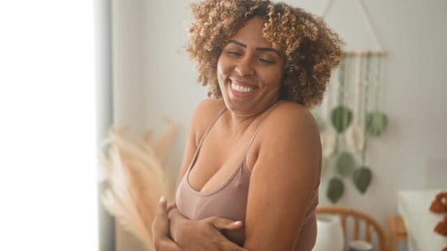Woman with bare shoulders and crossed arms hugging chest, smiling broadly in a cozy studio space with plants and chair; self love joy.