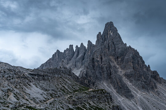 View of Paternkofel mountain peak with rugged rocky slopes, a winding hiking trail, and a dramatic cloudy sky in Auronzo di Cadore, Veneto, Italy.