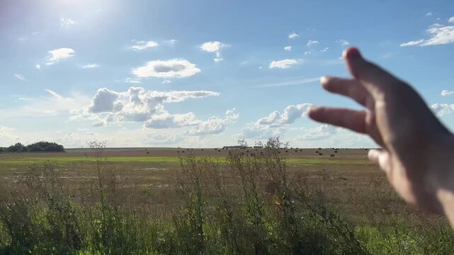 A view from a car window shows a woman s hand peeking out toward the horizon of a field dotted with sheaves of grain on a summer. A shot taken while driving down the road. Travel, vacation, packages