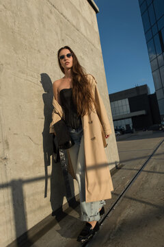Elegant stylish young woman in a beige trench coat, dark sunglasses, and jeans posing confidently near a concrete wall in a sunny city street. Modern urban fashion lifestyle concept.