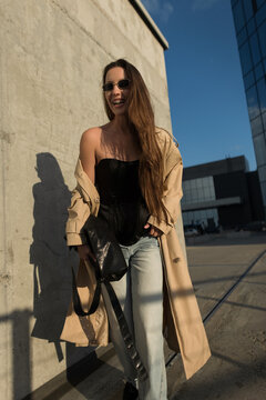 A cheerful young woman with braces, wearing a black corset, beige trench coat, and jeans, laughs joyfully while walking outdoors on a sunny city street with concrete and glass architecture.
