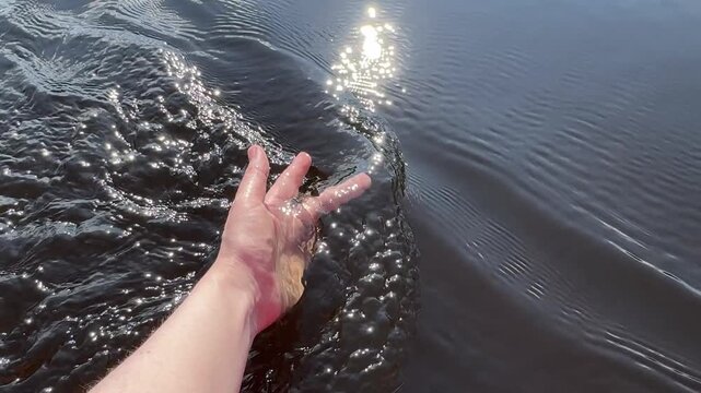 A close-up of a woman's hand dipped into the river, with waves reflecting the sun and glints of light during a river trip. Rafting, swimming, river trips, and relaxing on the river