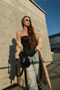 Low-angle view of a fashionable young woman in a beige trench coat and black corset, wearing sunglasses. She poses confidently in a modern urban concrete landscape under a clear blue sky.