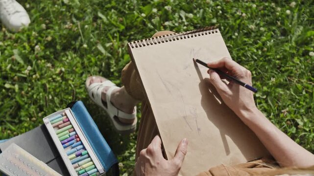 Unknown woman sketching on paper with pencil while sitting on grass. Footage is ideal for commercial use in mental wellbeing campaigns, art education, or hobby promotions