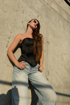 A low-angle shot of a stylish young woman leaning confidently against a rough concrete wall under direct sunlight. She is wearing a black bustier, wide-leg blue jeans, and sunglasses.