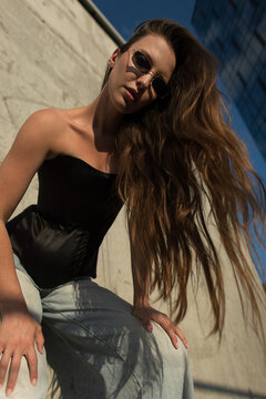 Stylish woman with long, flowing brown hair, black strapless corset, and jeans, posing confidently in an urban environment of concrete walls and glass skyscrapers. Pretty summer beauty girl
