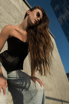Low-angle fashion portrait of a stylish young model with long wavy hair, sunglasses, black corset top, and jeans posing outdoors against a concrete wall on a sunny urban day.