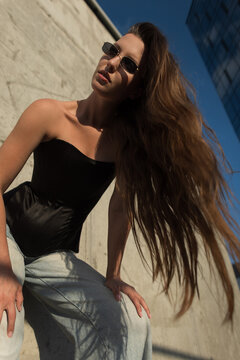 Stylish young woman with long flowing hair and sunglasses posing outdoors against a concrete wall. Low angle portrait of a trendy female model in a black corset top and jeans in sunny urban setting.