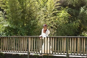 Fototapeta premium Young, brunette, Andalusian woman, beautiful, wearing a white flamenco dress and flowers in her hair, posing in the middle of a wooden bridge in a city park.