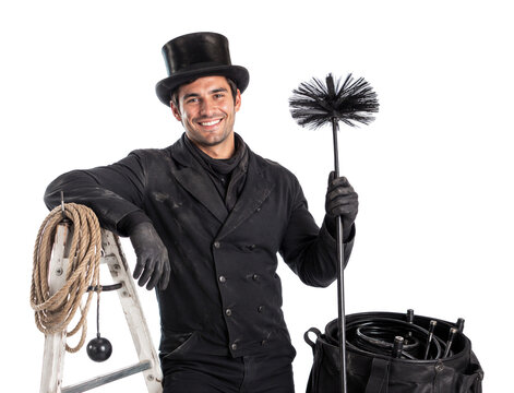 Young smiling chimney sweep in traditional black workwear holding a chimney brush beside ladder, rope and tools, professional cleaning service and skilled trade concept.