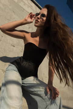 Stylish young woman in sunglasses and a black corset top, sitting on concrete steps in an urban setting. Low-angle sun-drenched fashion portrait with blowing hair. Light blue jeans. Confidence.