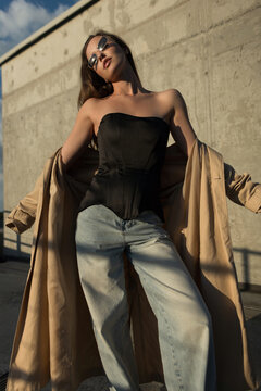 Confident female model in a black corset and jeans poses on a sunny city rooftop, dramatically holding a beige trench coat. Modern urban street style with sunglasses.