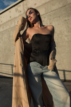 Stylish young woman in a black corset top, jeans, and beige trench coat posing outdoors against a concrete wall. Low angle portrait with sunglasses in warm sunlight.