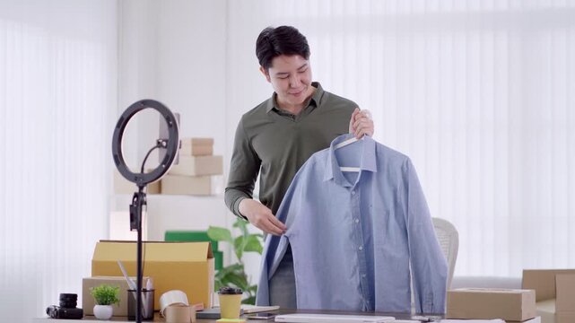 Young Asian male entrepreneur presenting blue shirt on hanger while filming product review for online store