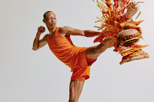 Male martial artist in orange training gear delivering high kick into exploding burgers and fries for fitness campaign background and health promotion banner