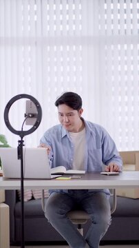 Young Asian entrepreneur writing notes during online selling session with laptop and smartphone in home studio