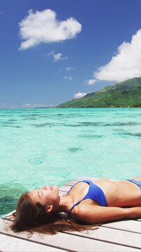 Bikini body sexy woman sunbathing on beach. Luxury travel girl tanning relaxing lying down on tropical getaway in swimsuit suntan. Raw unedited vertical video of real woman model without makeup