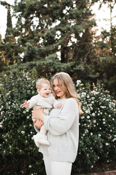 Cute woman holding baby wearing similar beige knitted sweaters over blooming bushes with flowers and sun light. Motherhood.