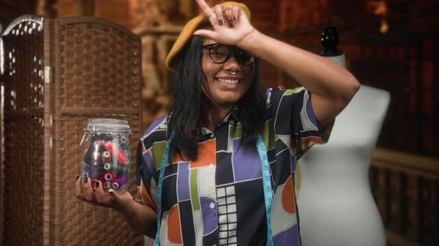 Woman holding jar of thread and spools with measuring tape around neck while making l sign on forehead in studio; creative joyful.