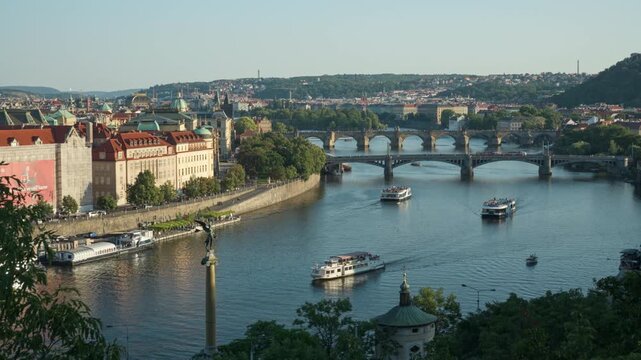 A scenic Prague cityscape showcases the Vltava River tourist cruise route. Famous city bridges frame the historic panorama under a summer sky. Passenger boats offer urban water tours.
