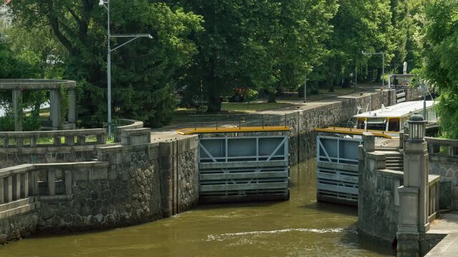 Metal lock gates open at the historic Smichov Lock Chamber in Prague. Heavy canal infrastructure regulates river transport for excursion boats. River navigation operates during summer season.