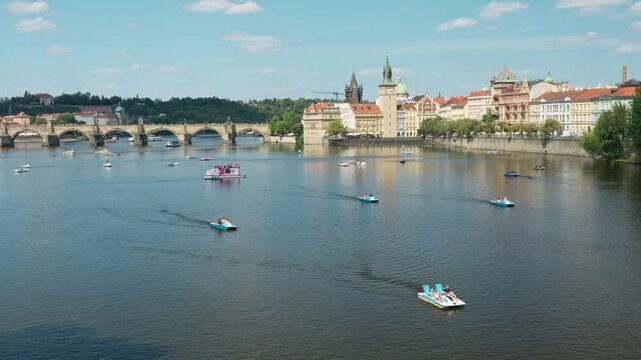 Tourists ride pedal boats on the Vltava River in historic Prague city. The famous Charles Bridge and traditional waterfront stand under a sunny blue sky. Popular European water recreation.