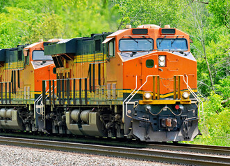 Two orange and black diesel‑electric train locomotives traveling through a green wooded corridor on a rural railroad line © dvande