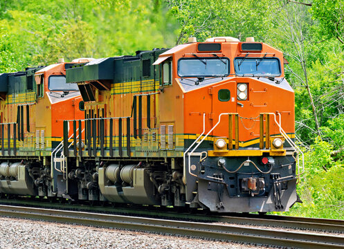 Two orange and black diesel‑electric train locomotives traveling through a green wooded corridor on a rural railroad line