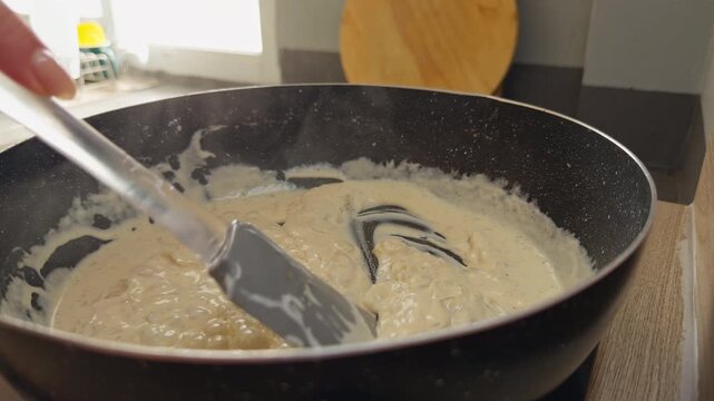 Hand Stirs Creamy Sauce in a Black Skillet on a Kitchen Counter, Showing Warm Food Preparation and Home Cooking Slow motion