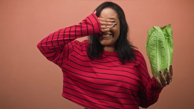 Young woman holding romaine lettuce while covering her eyes with hand in studio; playful healthy eating moment.