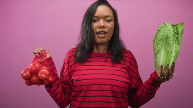 Woman holds romaine lettuce in one hand and a mesh bag of tomatoes in the other in a studio setting, facing forward and presenting both items; contemplation.