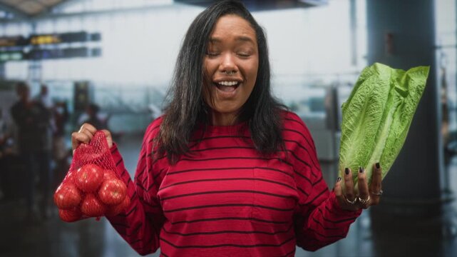 Young african american woman holds a net bag of tomatoes and romaine lettuce, arms extended and mouth open in an airport terminal; playful choice.