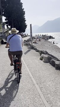 Man in blue shirt and straw hat cycling along Lake Garda in Malcesine, Italy
