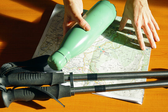 One hand lifts thermos from table while the other touches open paper map beside trekking poles. Reveals final route check, analog navigation, and last-minute preparation before heading out on foot.