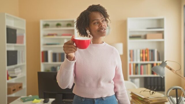 Young woman holding red coffee cup and smiling while standing by a desk with bookshelves, lamp and monitor in a studio; contentment calm morning.
