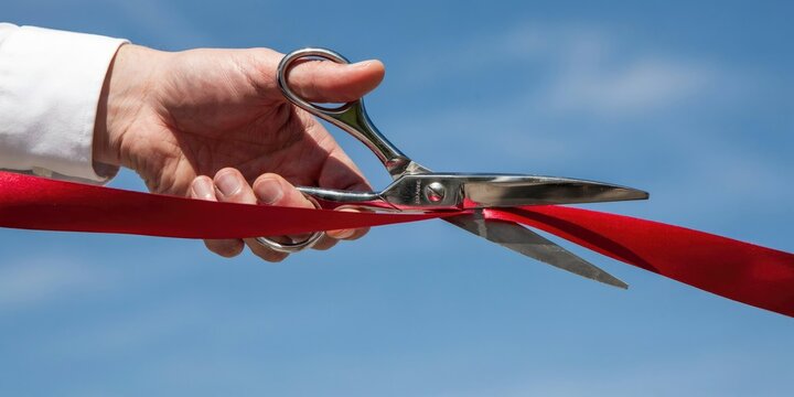 Hand cutting a red ribbon with silver scissors against a blue sky