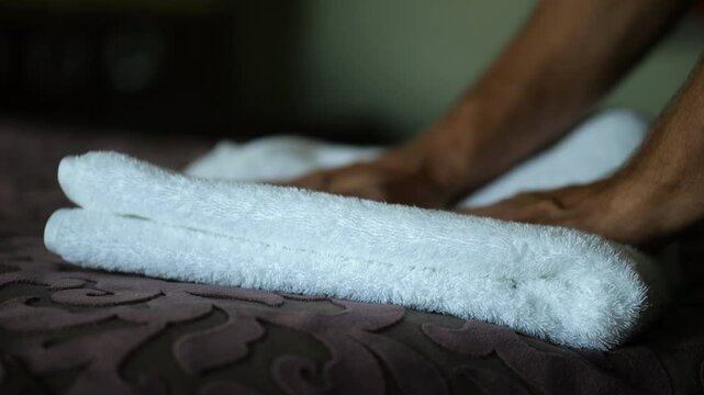 Male hands of housekeeping adjusting soft white terry towel on a neatly made bed in a modern hotel room. Service personnel preparing clean bedding and hospitality interior for guest accommodation