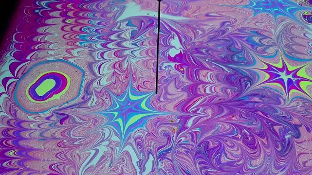 Artist's hand using a marbling tool to create swirling patterns in vibrant purple, pink, blue, and green ebru art on water, fluid abstract design, creative art technique.