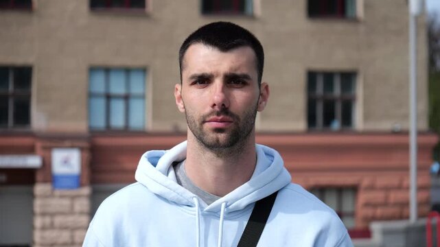 Portrait of young confident bearded man in casual clothing with cityscape background. Close up of handsome guy looks into camera standing at city environment at sunny day
