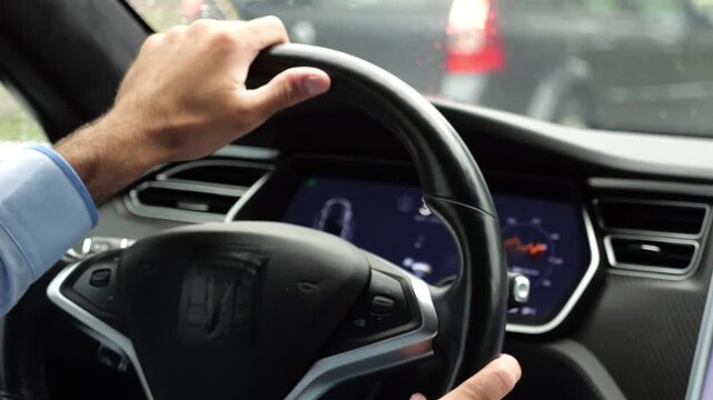 Male hands of businessman holds steering wheel while driving electrical vehicle at rain city. Young man commuting to work at rainy day. Businessperson operating electric car on urban road. Close up