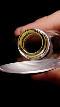 Vertical video. Hands Pour Thick Brown Liquid From a Glass Bottle Into a Metal Spoon Against a Black Background, Showing Careful Measurement and Preparation