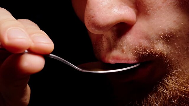 Bearded Man Tastes Brown Liquid From a Metal Spoon in Extreme Close Up Against Black Background, Showing Food Sampling and Flavor Evaluation